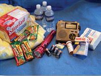 Una foto de los artículos esenciales de la bolsa de viaje. Incluye agua embotellada, batidos slimFast, barritas energéticas, una linterna, una radio, pilas, tiritas y un frasco de medicamentos. 