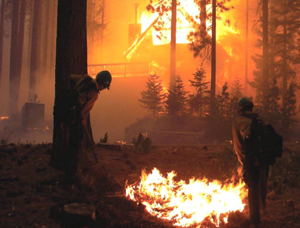 A picture of a home engulfed in flames with two fire fighters working to extinguish a spot fire in the foreground of the picture. 