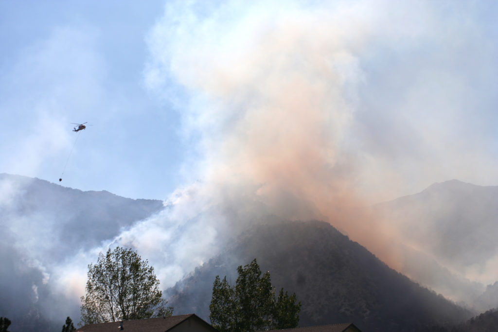 Picture of trees, mountains and smoke with a helicopter with water in the air. 